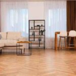 Modern living room with herringbone SPC flooring in Kerala, featuring a light beige sofa, brown curtains, and a minimalist black shelf.